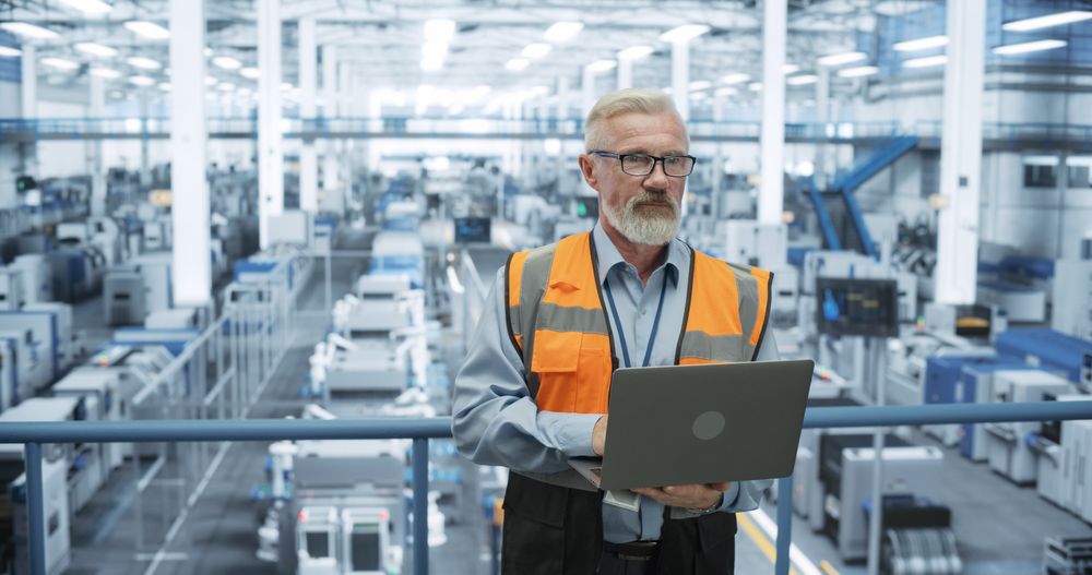 Experienced Male Engineer Standing on a Platform, Wearing a High-Visibility Vest, Using Laptop Computer and Overlooking Production at a Modern Automated Electronics Manufacture with Robotic Arms
