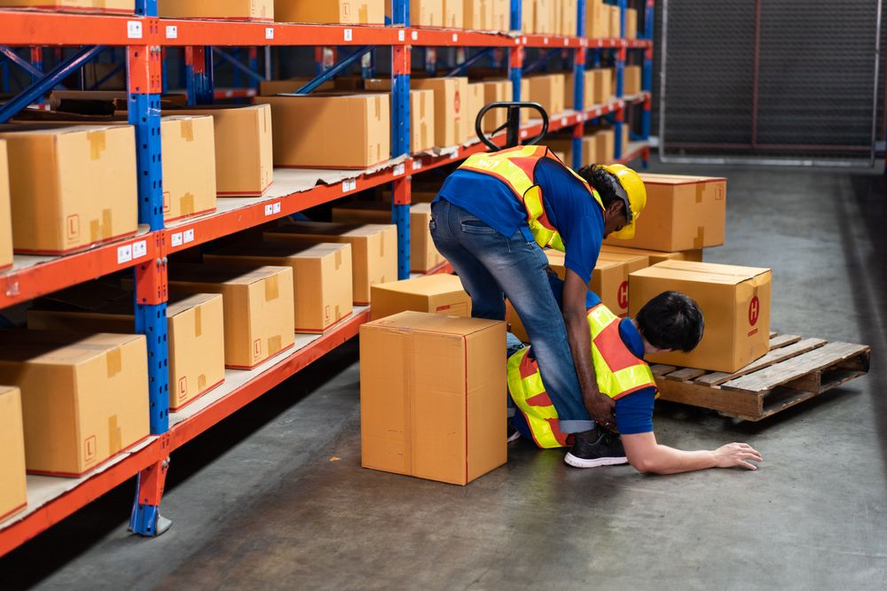 Workplace accident in a warehouse, a worker helping an injured colleague on the floor.