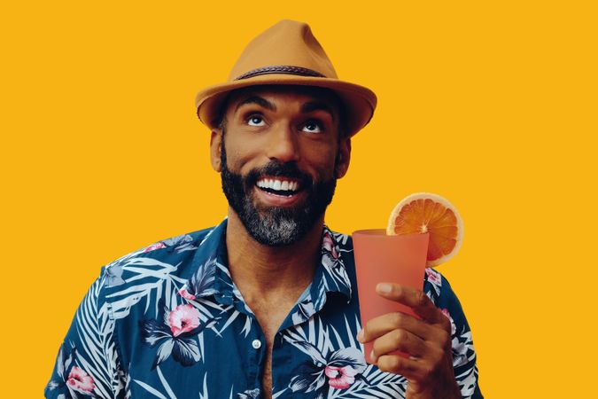 Happy male in hat and colorful shirt looking up while holding cocktail with yellow background