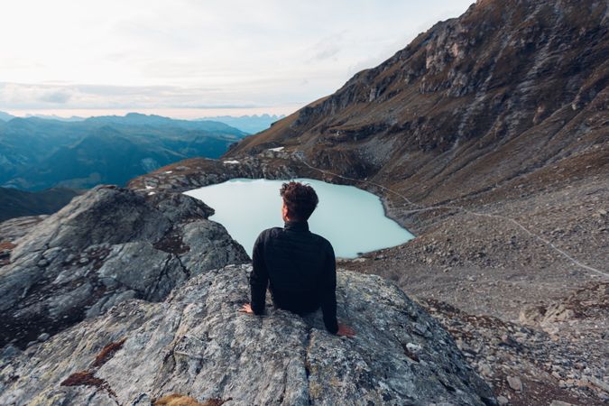 Back view of man in dark jacket sitting on water of frozen lake in mountain