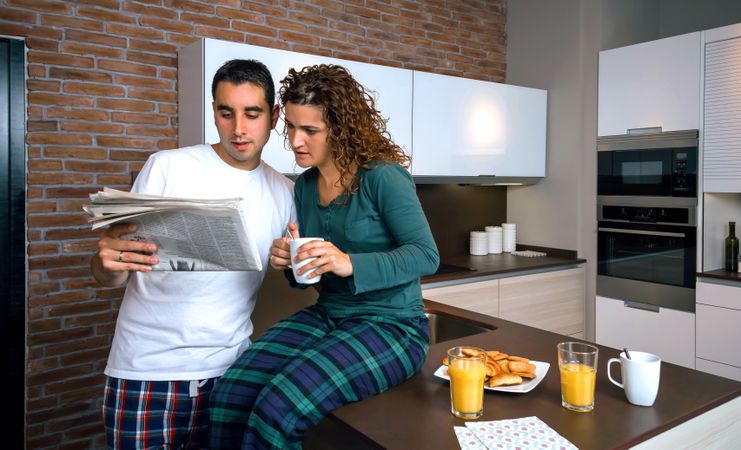 Couple in pajamas reading paper together in kitchen at breakfast time with coffee and orange juice