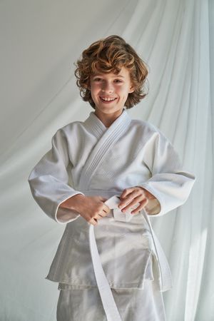 Positive preteen karate kid in kimono standing near curtain