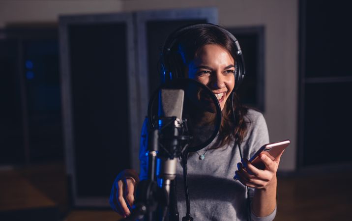 Smiling female playback singer with microphone and mobile phone in recording studio