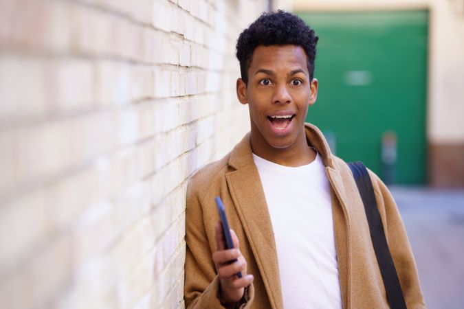 Surprised Cuban male leaning against wall holding his phone