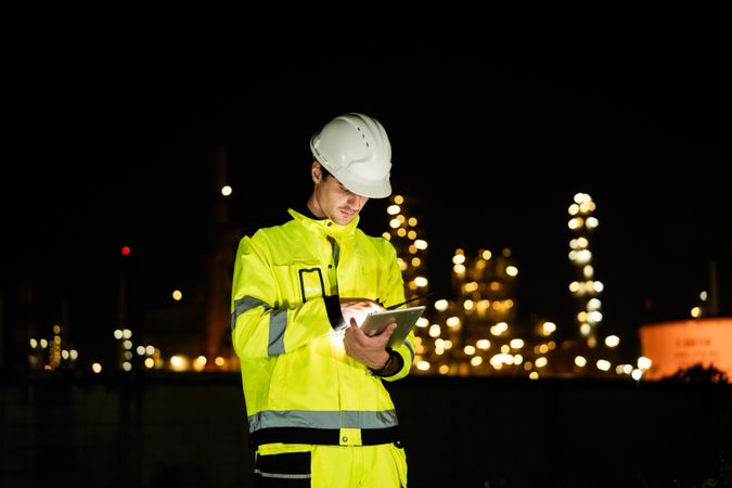Engineer worker watching work monitor on digital tablet and checking for quality control at construction site of petroleum chemical industry or refinery oil plant factory overtime at night
