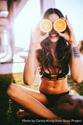 Young woman holds orange halves up to her eyes while sitting on the grass R5RYW0