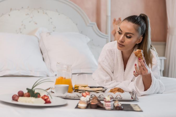 Woman in bathrobe laying forward and eating breakfast on bed