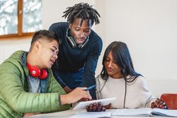 Three multi-cultural students engage in an interactive learning session with notes and digital devices, sharing knowledge