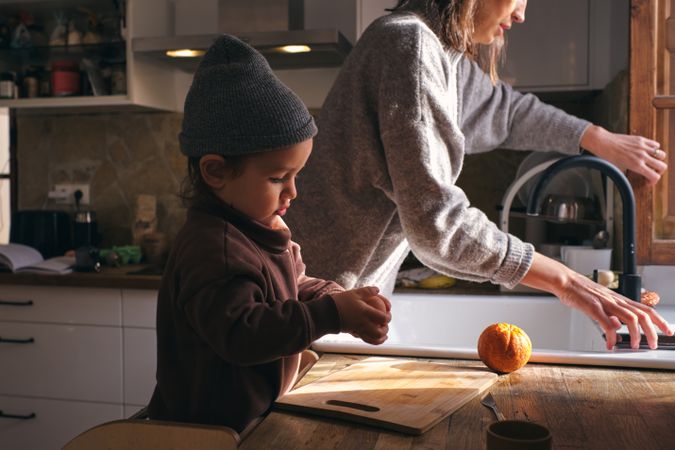 Calm little boy peeling oranges near mother washing dishes