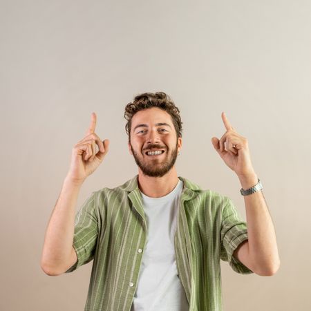 Happy young man pointing up with both fingers on a neutral background.