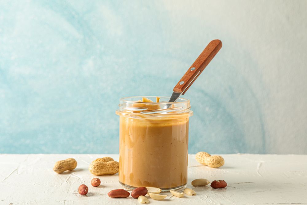 Glass jar with creamy peanut butter and spoon, and peanut on table against light background, space for text