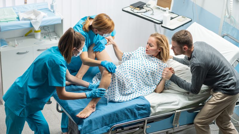 High Angle Shot of a Woman in Labor Pushes to Give Birth, Obstetricians Assisting, Spouse Holds Her Hand