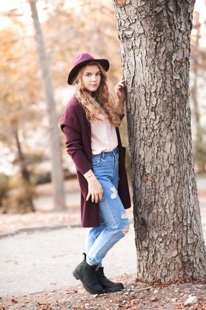 Neutral teenage girl in purple coat and hat standing beside tree