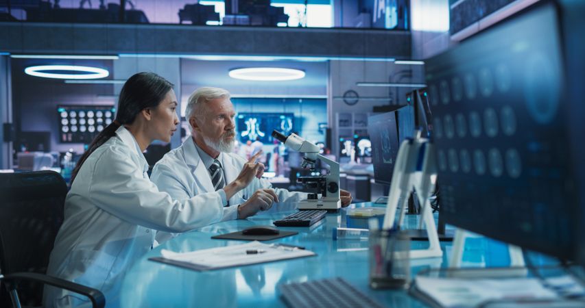 Biotechnology Medical Research Center: Female Asian Neuroscientist Using Desktop Computer And Talking To Male Surgeon With Microscope.
