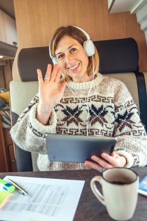 Female sitting in back of camper van waving with digital tablet, vertical