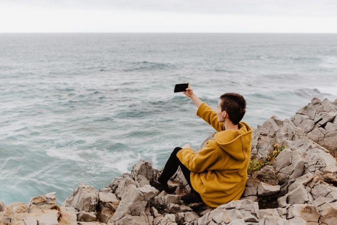 Woman in yellow hoodie taking selfie on smartphone on rocky shore