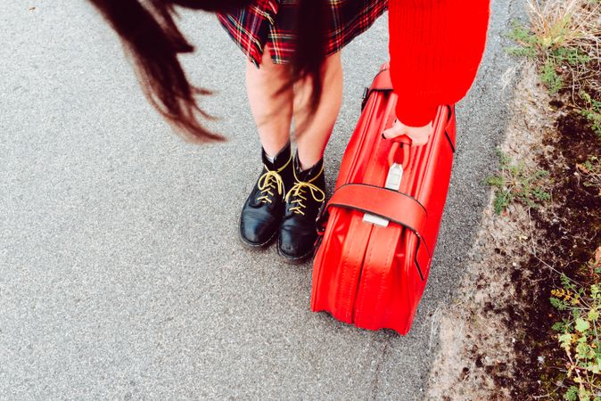 Woman in stylish boots with red suitcase standing on road