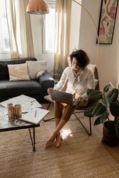 Woman with laptop on chair near low table with pencils, brushes and paper in room