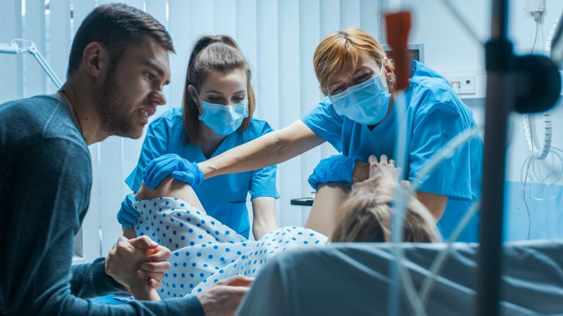 Emergency In the Hospital: Woman Giving Birth, Husband Holds Her Hand in Support, Obstetricians Assisting