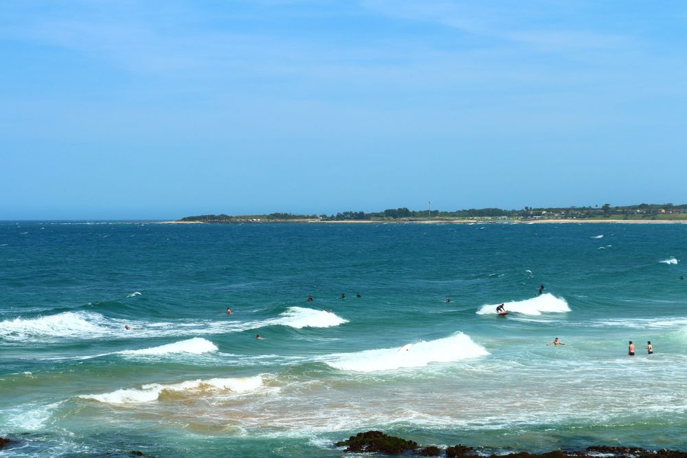 Swimmers Enjoying The Waves Of The Blue Ocean Near A Rocky Shore On A Clear Sunny Day