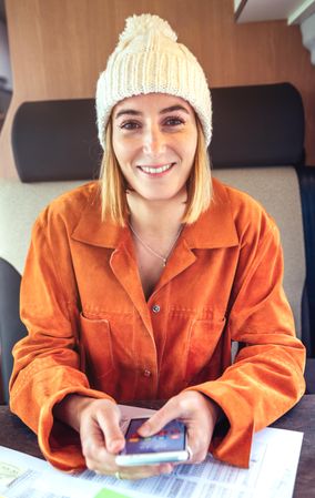 Woman in orange shirt sitting and smiling in van with smartphone, vertical