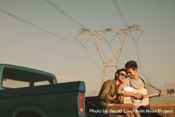 Young woman and man sitting on edge of pickup truck playing guitar while parked on country road 5wlXR4