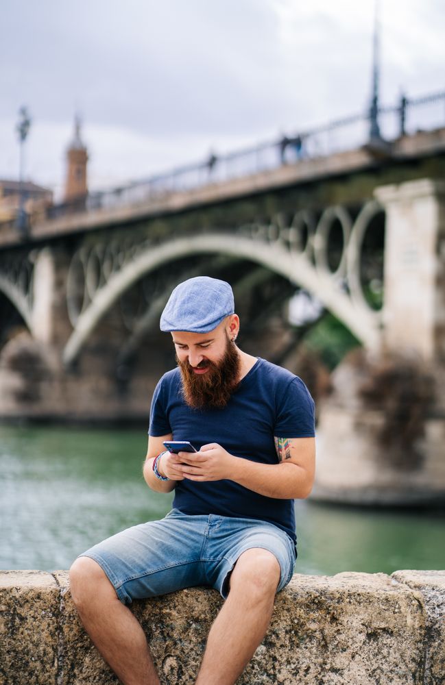 Bearded young man in casual wear using smartphone while walking on quay