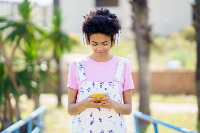 Content female in floral overalls and headphones texting on phone outside