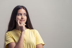 Thoughtful teenage girl touching face with finger while looking away on neutral background