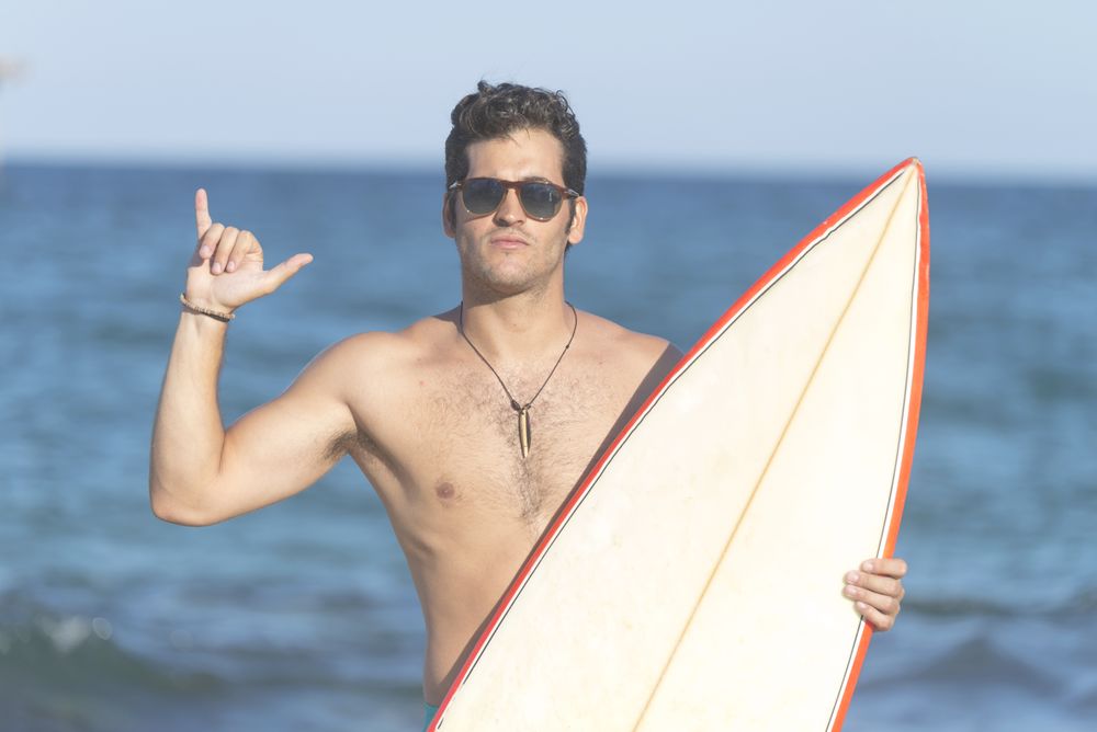 Male surfer standing with red outlined board near sea making the “hang loose” sign