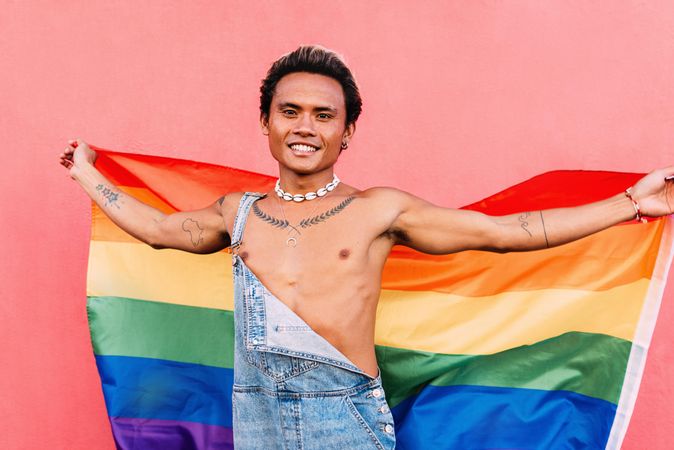 Happy male in open overalls looking at camera and holding a pride flag with his arms wide