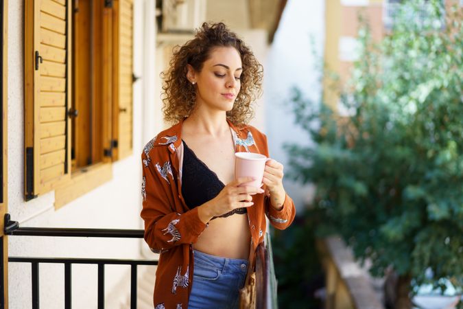 Young stylish smiling woman with cup of coffee standing on balcony at home