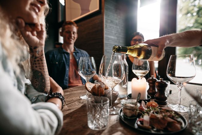 Man hand pouring white wine from the bottle into glasses with friends sitting around the table
