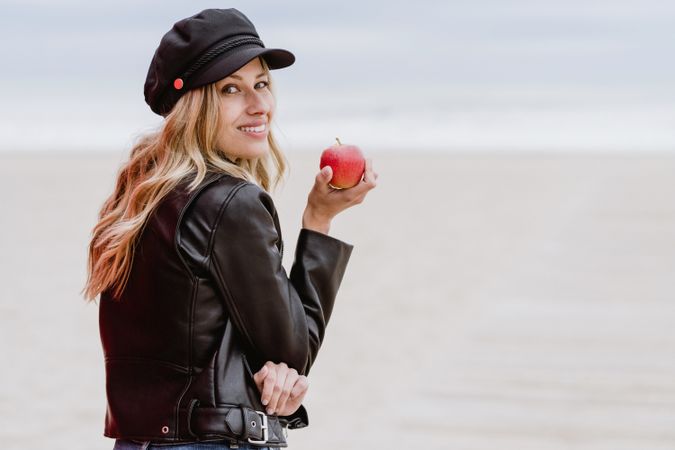 Smiling woman with apple looking at camera over shoulder standing on beach