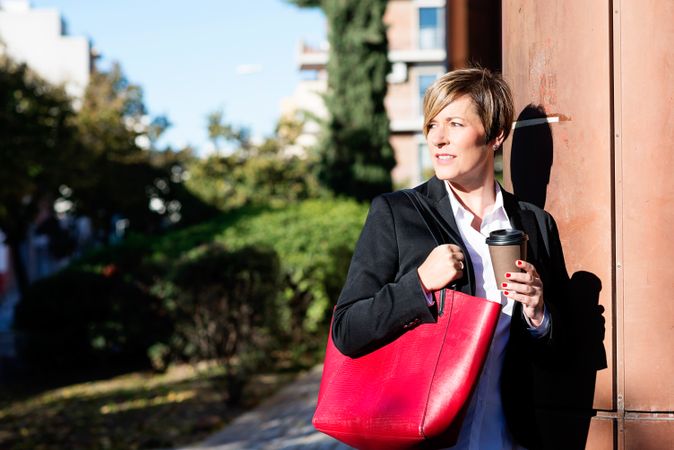 Female in blazer standing in the sun with coffee and large red handbag
