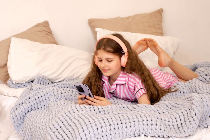 Girl in pink pajamas lying on bed using smartphone.
