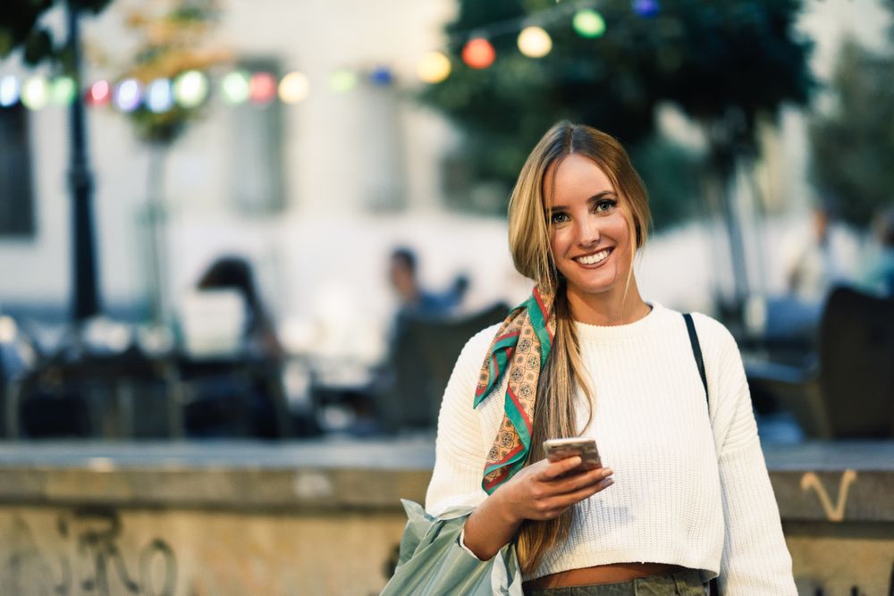 Chic woman with scarf in hair smiling outside holding phone