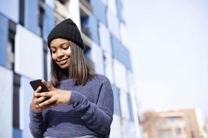 Female in wool hat and blue sweater checking smart phone outside blue building with space for text