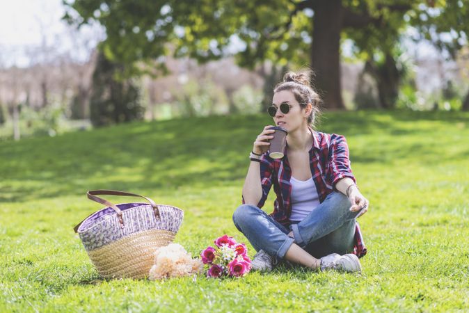 Front view of a young hipster woman sitting on grass in a park while holding a take away mug in a sunny day