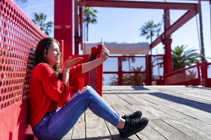 Side view of smiling woman in red sitting taking selfie on mobile phone outdoors on a bright day