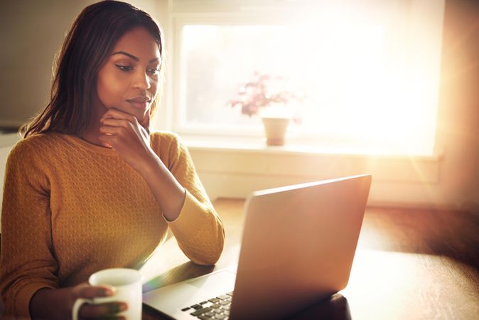 Contemplative Black woman on laptop in sunny room