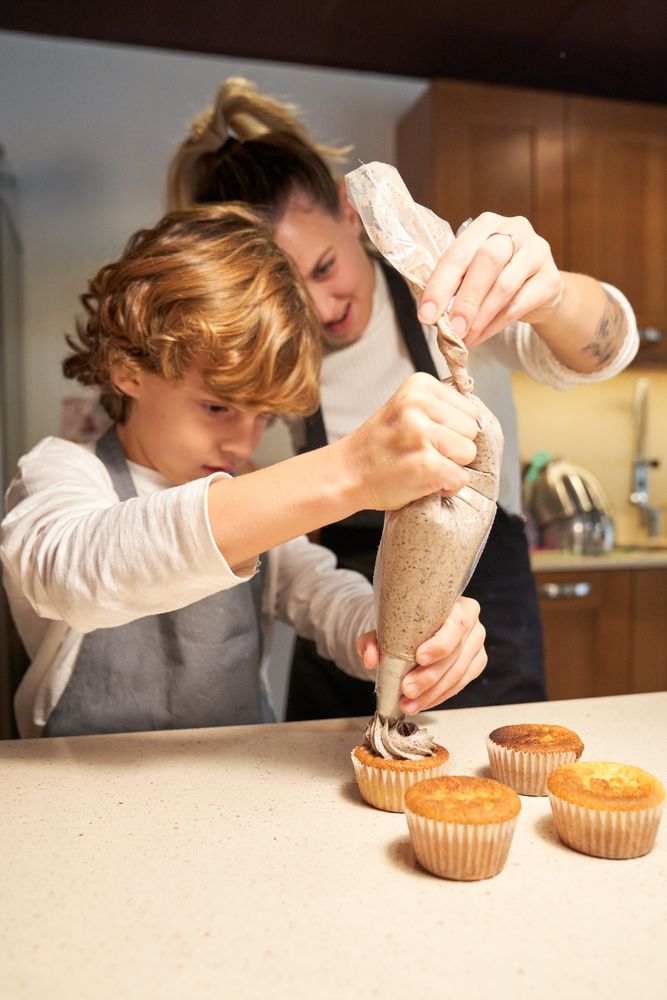 Vertical photo of a girl wearing an apron helping a boy to decorate cupcakes with a pastry bag