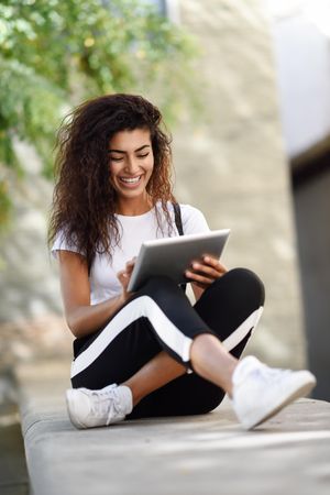 Happy Arab woman looking down at her digital tablet while sitting outdoors, vertical