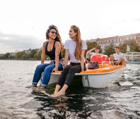 Two women friends sitting in front pedal boat with feet in water and man in background
