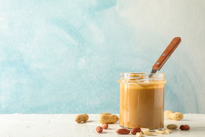 Glass jar with creamy peanut butter and spoon, and peanut on table against light background, space for text