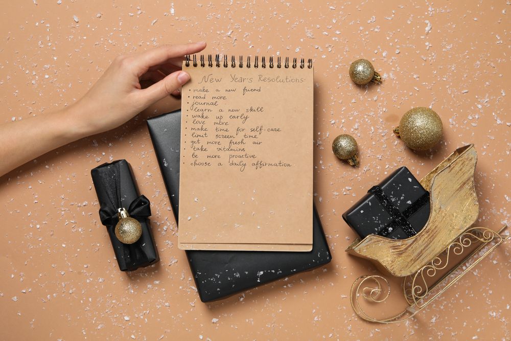 Gift boxes, decorations and notepad with list in hand on beige background, top view