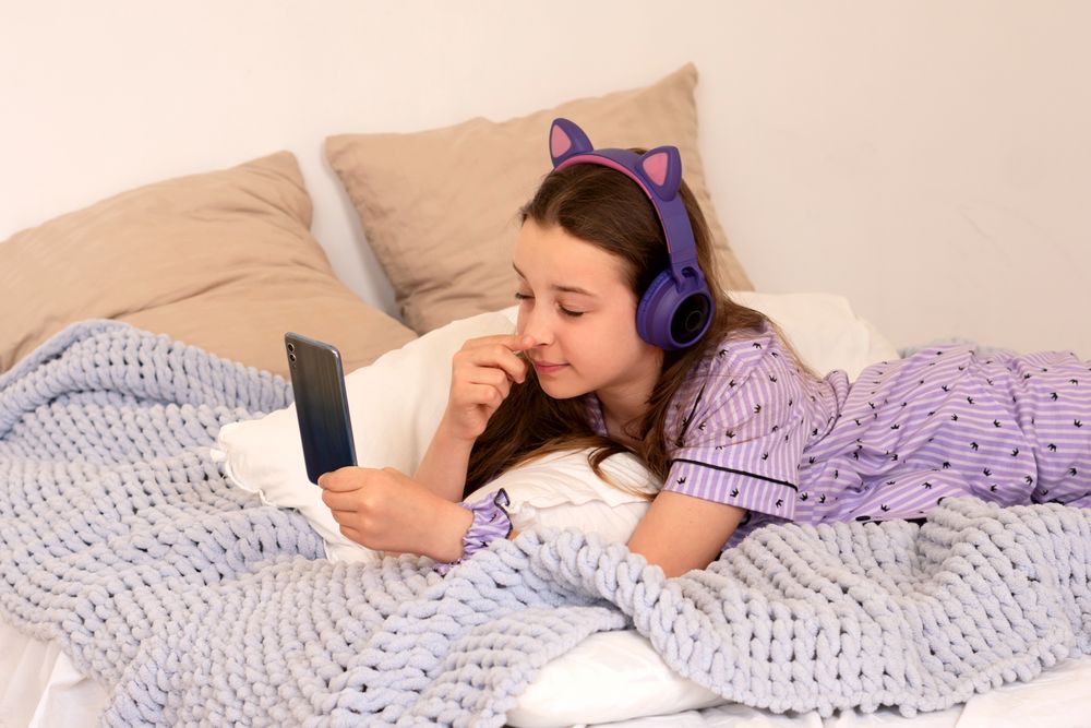 Girl in bed with headphones, browsing smartphone.