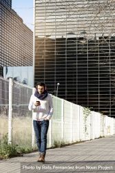 Young man in scarf walking past metallic fence and holding smartphone outdoors 5oD19k