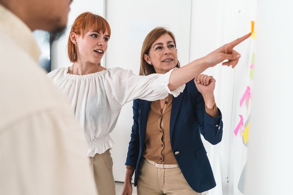 A woman explains her project to colleagues by pointing at a colorful sticky note on a diagram on the wall