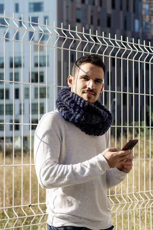 Young man looking up wearing scarf leaning on metallic fence and holding smartphone outdoors